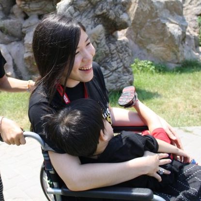 Side pic of Xian smiling sitting in wheelchair and holding a child on her lap
